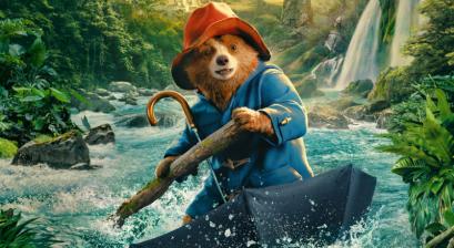 Plakat filmu animowanego, na pierwszym planie miś paddington w czerwonym kapeluszu i pelerynie przeciwdeszczowej płynący po rwącej rzecze między górami