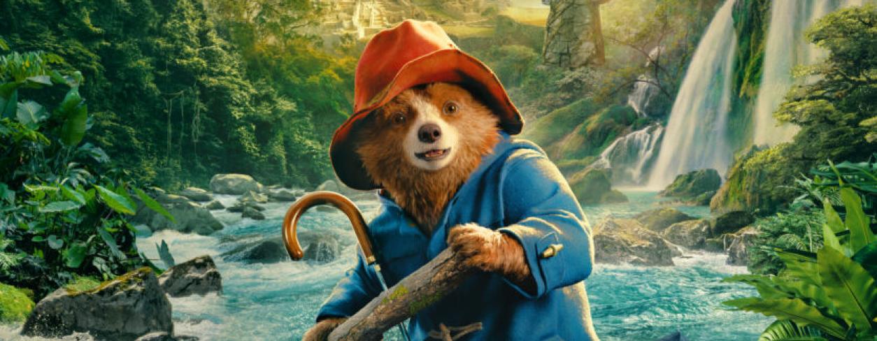 Plakat filmu animowanego, na pierwszym planie miś paddington w czerwonym kapeluszu i pelerynie przeciwdeszczowej płynący po rwącej rzecze między górami