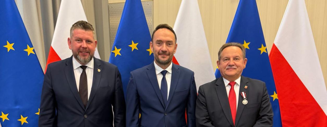 Burmistrz oraz osoby wręczające na tle flag 