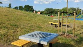 widok na nowo posadzone drzewa między stolikami do rozgrywek szachowych, na drugim planie widać plac zabaw znajdujący się przy zalewie Droniowickim