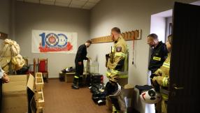Pomieszczenie w siedzibie ochotniczej straży pożarnej, z kartonami z nowym umundurowaniem stojący strażak po prawej stronie ubrany w nowy mundur, w kolorze khaki spodnie i kurtka -ognioodporne z kaskiem w ręce