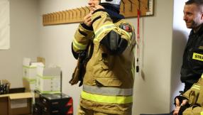 Pomieszczenie w siedzibie ochotniczej straży pożarnej, z kartonami z nowym umundurowaniem stojący strażak po prawej stronie ubrany w nowy mundur, w kolorze khaki spodnie i kurtka -ognioodporne z kaskiem w ręce